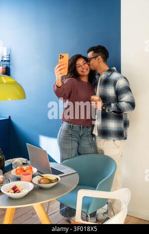 Ein lateinisches Paar macht ein fröhliches Selfie zu Hause in einer modernen Küche, lächelt, während der Mann der Frau die Wange küsst. Gemütlicher Lifestyle-Moment mit Laptop, c Stockfoto