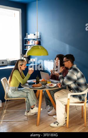 Drei multiethnische Mitbewohner teilen sich Frühstück und Kaffee an einem kleinen Esstisch in einer gemütlichen Küche, unterhalten sich und benutzen einen Laptop im Morgenlicht. Stockfoto