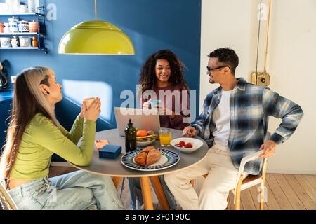 Multiethnische Mitbewohner teilen Frühstück und Kaffee an einem Küchentisch, plaudern und lächeln in der Nähe eines Laptops in einem hellen Apartment und genießen einen entspannten Morgen Stockfoto