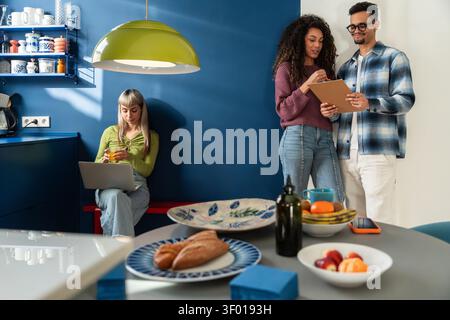 Multiethnische Mitbewohner in einer modernen Küche teilen sich einen entspannten Morgen, checken Notizen und ein Tablet, während man an einem Laptop arbeitet, mit Frühstück auf dem Tisch Stockfoto