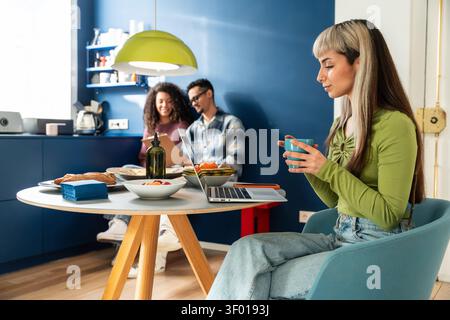Multiethnische Mitbewohner teilen sich einen ruhigen Morgen in einer modernen Apartmentküche, trinken Kaffee, plaudern und benutzen Laptops an einem kleinen Tisch mit Frühstück Stockfoto