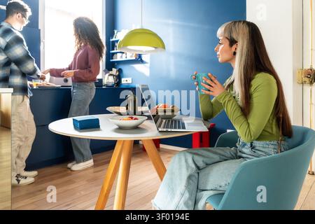 Multiethnische Mitbewohner teilen sich einen ruhigen Morgen in einer modernen Küche: Einer schlürft Kaffee am Laptop am Tisch, während zwei gemeinsam das Frühstück in einem bri zubereiten Stockfoto