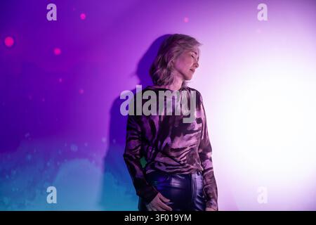 Futuristische ältere asiatische Frau in stimmungsvollem lila Neonlicht, im Profil vor einer leuchtenden Wand mit Bokeh-Punkten, in stilvollem Oberteil und Leine Stockfoto