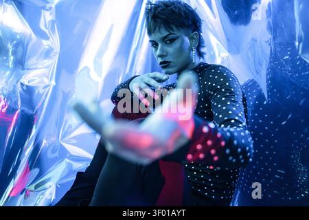 Cyberpunk-junge Frau posiert in einem neonbeleuchteten Studio, aus einem niedrigen Winkel aufgenommen, mit der Hand zur Kamera gerichtet, dramatische blaue und magentafarbene Beleuchtung, reflektierend Stockfoto