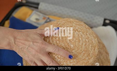 Frau, die einen Hartschalenkoffer packt, Hand mit blauen Nägeln mit Strohhut und sichtbaren Dollar auf dem Bett im Gebäude; Vorausschau. Stockfoto