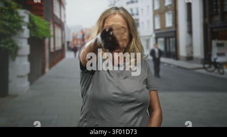 Eine ältere Frau mit blondem Haar zeigt den Finger direkt auf die Kamera auf einer städtischen Straße, die von Gebäuden und Fahrrädern gesäumt ist; Selbstvertrauen. Stockfoto