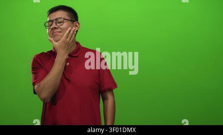 Junger Mann mit Hand auf dem Kinn, der in grünem Hintergrund im Studio posiert, mit rotem Polo und Brille; nachdenkliche Reflexion. Stockfoto