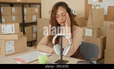 Junge hispanische Frau mit Hand zum Tempel, die Paketpapiere neben gestapelten Versandkartons im Gebäude liest; Stressbestandsmanagement. Stockfoto