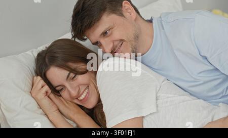 Frau lächelt auf dem Bett mit Hand auf Kissen, während der Mann ihre Taille berührt, Ringe sichtbar, entspanntes junges Paar, das sich auf weißen Kissen in einem Gebäude stützt; inti Stockfoto