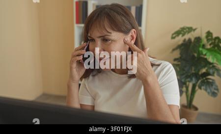 Frau, die auf dem Smartphone spricht, zeigt mit dem Finger auf den Tempel im Büro, wobei der Computerbildschirm sichtbar ist; nachdenklicher Fokus. Stockfoto