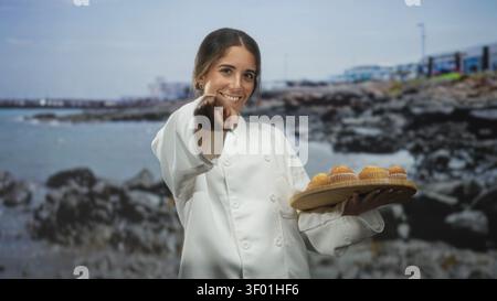 Die Chefin hält ein hölzernes Tablett mit Muffins und zeigt den Finger auf die Kamera auf der Straße an der felsigen Küste mit sichtbarem Pier; freundliche Gastfreundschaft. Stockfoto