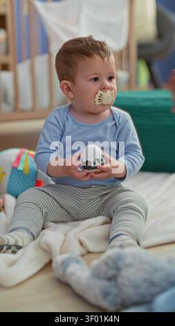 Junge, der Spielzeugball mit Schnuller hält, während er in einem Kinderzimmer in einem Gebäude sitzt; Neugier auf Zufriedenheit. Stockfoto