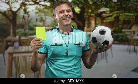 Mann Schiedsrichter hält gelbe Karte und Fußball, trägt Pfeifkopfhörer in Straßencafés; starke Autorität. Stockfoto