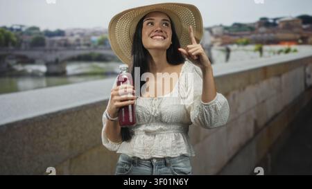 Junge Frau, die einen roten Saft in der Flasche hält und mit dem Zeigefinger auf einer Steinbrücke über einem Stadtfluss lächelt; glückliche Entspannung. Stockfoto