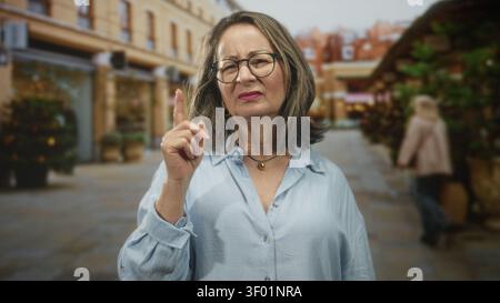 Hispanische Seniorin mit grauem Haar und Brille zeigt mit dem Zeigefinger und blinzelt, während sie in einer Straße steht, die von Geschäften und Pflanzern gesäumt ist; Missbilligung Stockfoto