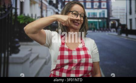 Die ranghöchste hispanische Frau zeigt Finger an Nase, während sie rote karierte Schürze und Brille auf der Stadtstraße in der Nähe der Treppen des Stadthauses trägt; verspielte Wärme. Stockfoto