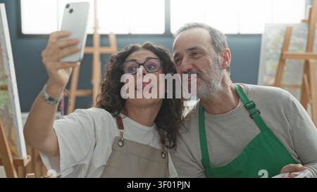 Eine mittelgroße Frau hält ein Smartphone für Selfie, während ein Mann in grüner Schürze im Kunststudio die Zunge herausragt; verspielt. Stockfoto