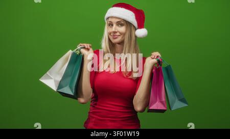 Frau mit mehreren Einkaufstaschen in beiden Händen, lächelt mit dem Kopf und trägt einen weihnachtsmann-Hut vor grünem Hintergrund in einem Studio; Urlaub Stockfoto
