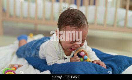 Junge weinend mit offenem Mund, der bunte Rassel auf blauem Kissen auf dem Bett im Gebäude hält; Traurigkeit Verletzlichkeit. Stockfoto