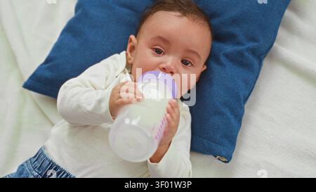 Hispanischer Junge, der weint und eine Milchflasche an den Mund hält, auf einem blauen Kissen in einer Futterszene im Studio, mit sichtbaren Tränen und geballter Hand; Hunge Stockfoto
