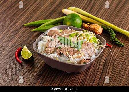 Pho bo serviert in einer Keramikschale mit Reisnudeln Rindsscheiben Sojasprossen Basilikum Limette Chili auf dunkler Holzoberfläche. Warmes natürliches Licht hebt frische Akzente Stockfoto