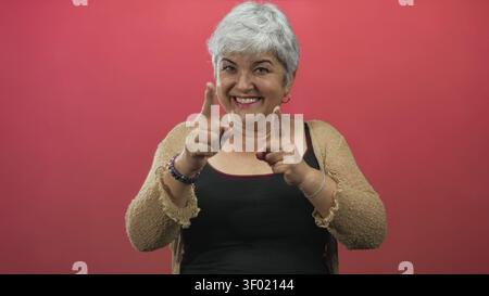 Frau, die mit einem Lächeln auf beide Hände zeigt, eine Strickjacke und ein schwarzes Tanktop vor einem roten Hintergrund trägt; verspieltes Selbstvertrauen. Stockfoto