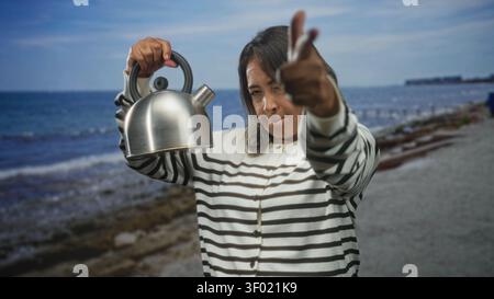 Die hispanische Frau hält einen Metallkessel und zeigt den Finger zur Kamera in einem Studio mit Strandkulisse; Selbstvertrauen. Stockfoto