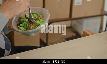 Mann isst Salat aus Plastikbehältern, hält Gabel an Mund neben Pappkartons im Gebäude; zufriedene kleine Unternehmen. Stockfoto