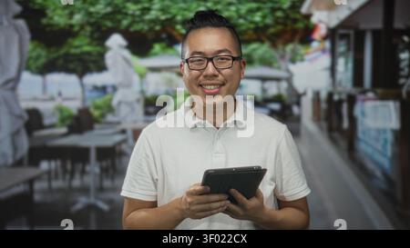 Mann, chinesisch und jung, lächelnd und klopfend mit beiden Händen auf einer Restaurantterrasse an der Straße, während er Brille und ein weißes Polo trägt; CAL Stockfoto