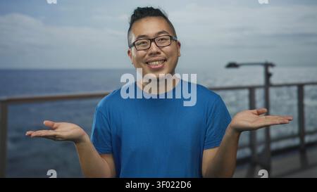 Mann in blauem T-Shirt mit Brille und Handzucken auf Kreuzfahrtschiff Deck am Geländer und Meer; Unsicherheit Neugier Offenheit. Stockfoto