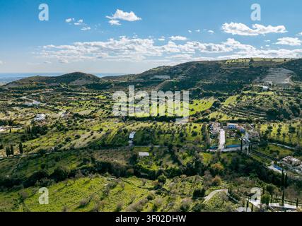 Blick aus der Vogelperspektive auf das historische Dorf Pano Lefkara, eingebettet in sanfte grüne Hügel, mit Terrassenfeldern, verstreuten Bäumen und einem hellblauen Himmel Stockfoto