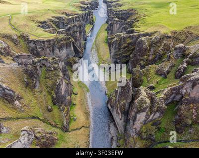 Blick aus der Vogelperspektive auf den tiefen Fjadrargljufur Canyon mit einem Fluss, der durch steile, moosbedeckte Felsformationen in Island fließt. Stockfoto
