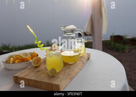 Eine erfrischende Limonade wird auf einer Terrasse serviert, die mit einem hölzernen Servierbrett und einer Keramikschale ausgestattet ist. Die Nahaufnahme zeigt zwei Gläser gefüllt mit Limonade und Strohhalmen, Setp. Stockfoto