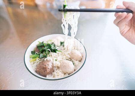 Thailändische Nudelsuppe mit Fleischbällchen und Grünzeug, serviert in einer weißen Schüssel mit Stäbchen, die sie hochheben Stockfoto