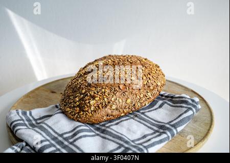Frisch gebackenes mehrkörniges Brot auf einem runden Holzbrett mit einem blau karierten Küchentuch vor einem hellen weißen Hintergrund, ein gesundes Brot Stockfoto
