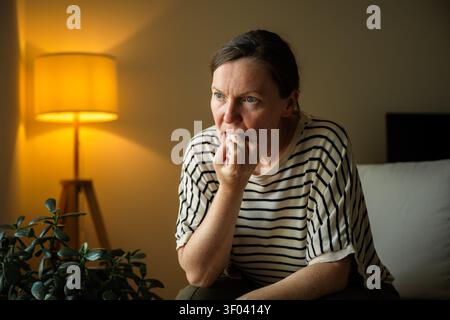 Eine Frau mittleren Alters beißt nervös in die Nägel, während sie abends zu Hause sitzt. Angst, Stress, Unsicherheit und emotionales Belastungskonzept. Wählen Sie Stockfoto