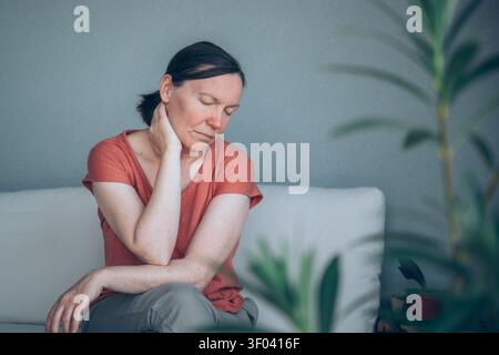 Reife Frau, die unter starken Nackenschmerzen leidet, während sie auf dem Balkonsofa sitzt und durch Oleanderblätter gesehen wird. Muskuloskelettale Beschwerden und Gesundheit Proble Stockfoto