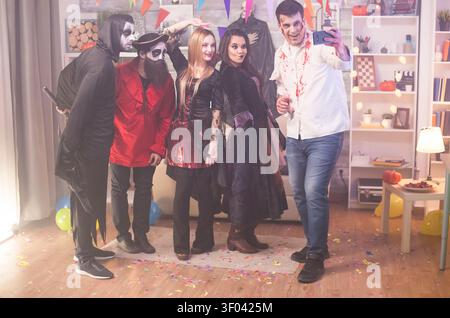 Der junge Mann und seine Freunde machen ein Selfie auf einer Halloween-Party, während sie Kostüme tragen, und teilen einen fröhlichen Moment mit theatralischem Design, dunkler Atmosphäre und klaren Kostümcharakterdetails. Stockfoto