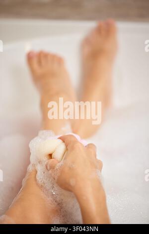 Frau schrubbt ihre Beine mit einem Schwamm, erzeugt Seifenblasen und entspannt sich in einer warmen Badewanne für persönliche Hygiene und Selbstpflege Stockfoto