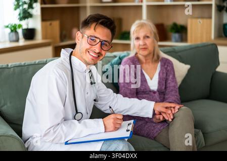 Ein freundlicher Arzt lächelt, während er sich während eines Hausbesuchs Notizen macht und sanft die Hand eines älteren Patienten hält, um beruhigend zu sein und Stockfoto