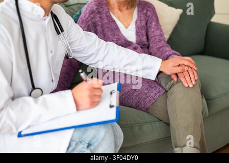 Ein Arzt zeichnet medizinische Informationen auf einem Klemmbrett auf und bietet gleichzeitig Komfort für eine ältere Patientin in ihrem Wohnzimmer. Stockfoto