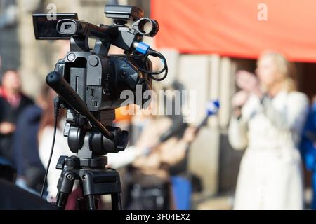 Nahaufnahme einer professionellen Videokamera, die ein Live-Interview im Freien aufzeichnet, verschwommener Reporter und Mikrofon im Hintergrund, repräsentierend die Medienberichterstattung, j Stockfoto