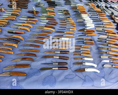 XALO, SPANIEN - 20. JUNI 2026. Zahlreiche Klappmesser mit verschiedenen Holz- und Kunststoffgriffen sind auf einem blauen Tisch auf einem Straßenmarkt aufgestellt. Stockfoto