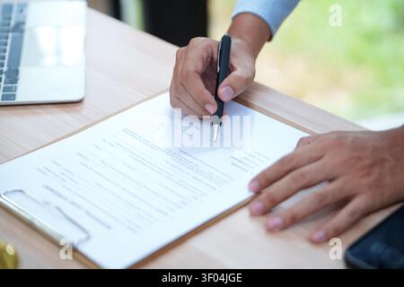 Mann unterzeichnet Dokument in Zwischenablage mit Laptop im Hintergrund für Stockfoto