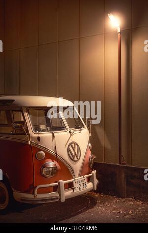 In einer ruhigen Seitenstraße in Kyoto, Japan, steht ein Vintage-Van des Typs Volkswagen T1, dessen rot-weißes Gehäuse das warme Leuchten einer Straßenlaterne einfängt. Stockfoto