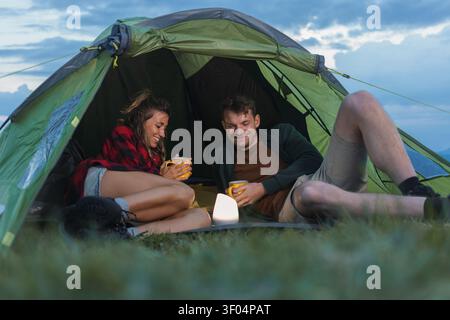 Zwei Wanderer entspannen sich in einem Zelt und genießen warme Getränke nach einem Tag der Wanderung in den Bergen in der Abenddämmerung. Stockfoto
