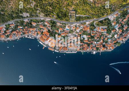 Ein geradliniges Drohnenfoto fängt die Küstenstadt Perast in Kotor Bay, Montenegro ein und zeigt rot gekachelte Dächer, enge Gassen und Boote entlang der tiefblauen Adriaküste. Der üppig grüne Hügel erhebt sich über die Stadt, mit einer gewundenen Straße A Stockfoto
