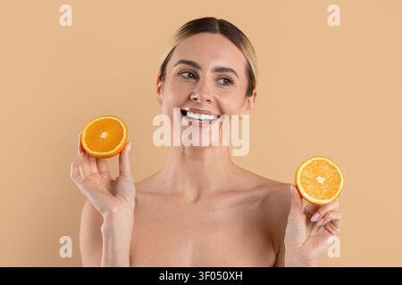 Junge schöne Frau lächelt, während sie frische Orangenhälften auf beigem Hintergrund hält Stockfoto