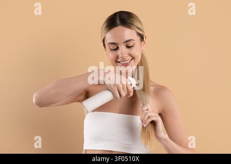 Junge schöne Frau lächelt, während sie Haarpflegeprodukt auf ihren Pferdeschwanz sprüht Stockfoto