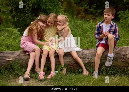 Drei Mädchen, die sich umarmen, während der Junge im Sommercamp im Park auf umgestürzten Bäumen sitzt. Stockfoto